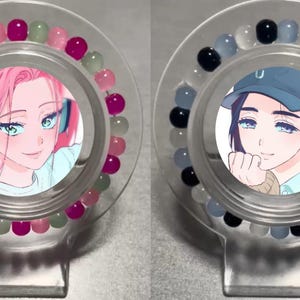 May include: Two clear plastic bracelets with colorful beads and a round image of a person inside. The bracelet on the left has pink, green, and white beads. The bracelet on the right has black, white, and gray beads.