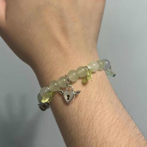 Pu&ograve; includere: Un braccialetto di perline verdi e grigie con un ciondolo a forma di cuore argentato con un buco della serratura. Il ciondolo ha piccole ali su entrambi i lati.