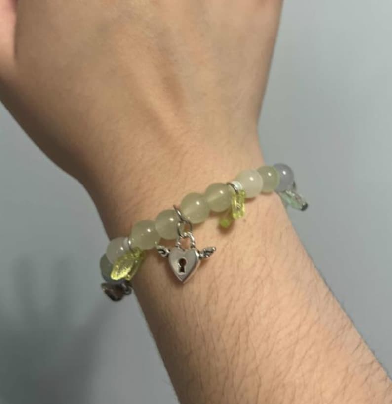 Pu&ograve; includere: Un braccialetto di perline verdi e grigie con un ciondolo a forma di cuore argentato con un buco della serratura. Il ciondolo ha piccole ali su entrambi i lati.
