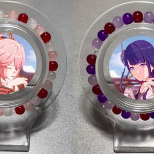 May include: Two clear plastic display stands with red and white beads on one and purple and white beads on the other. Each stand has a round image of an anime character inside.