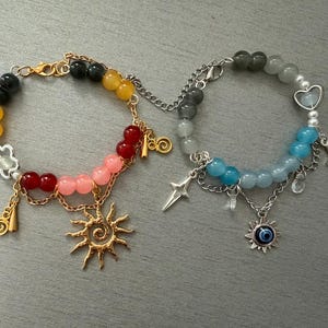 May include: Two linked bracelets featuring colorful beads and metal charms. The left bracelet has black, yellow, red, and pink beads with a gold sun charm. The right bracelet has grey, blue, and white beads with a silver star and evil eye charm.