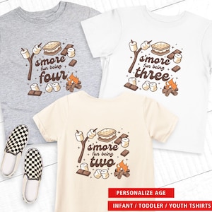 May include: Three children's t-shirts in gray, white, and cream, each with a s'mores graphic and age-specific text: "S'more fun being four," "S'more fun being three," and "S'more fun being two." The image also includes a pair of black and white checkered slip-on shoes. Text at the bottom reads "PERSONALIZE AGE" and "INFANT / TODDLER / YOUTH TSHIRTS."