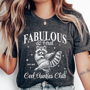 May include: Dark grey heather t-shirt with the text "FABULOUS & FERAL" and a raccoon graphic. The shirt also features the text "Cool Aunties Club" and "EST 2025".