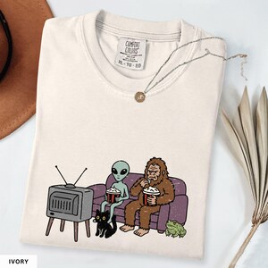 May include: An ivory t-shirt featuring a graphic print of a grey alien and a brown Bigfoot sitting on a purple couch eating popcorn while watching a vintage television. A black cat and a green frog sit on the floor in front of the couch.