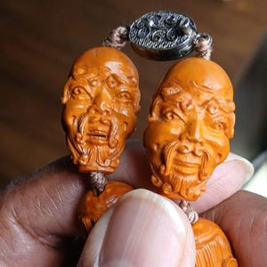 May include: Close-up of a carved wooden bead bracelet featuring detailed faces with beards. The beads are a warm orange-brown color, strung together with a tan cord, and connected by a silver clasp. The bracelet is held in a hand.