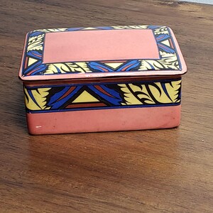 May include: A pink ceramic box with a geometric design in yellow, blue, and red. The box has a lid with a flat top.