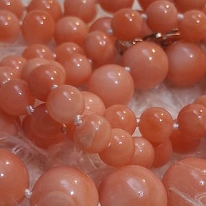 May include: A necklace composed of smooth, round, salmon-pink beads of varying sizes. The beads are strung together on a thin white cord, creating a multi-layered appearance with a polished finish that reflects light across the spherical surfaces.
