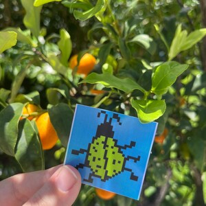 May include: A square sticker with a pixelated green ladybug design on a blue background. The sticker is held in front of a tree with green leaves and orange fruit.