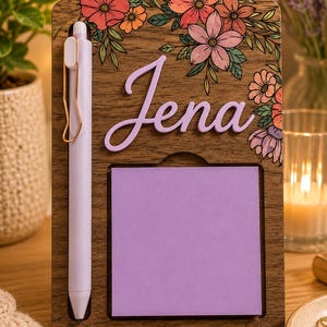 May include: Wooden desk organizer with a floral design and the name "Jena" in lavender. Includes a lavender pen and a square of purple sticky notes. The organizer is decorated with pink, orange, and purple flowers.