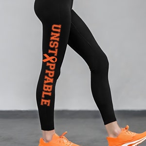 Może przedstawiać: Czarne legginsy z pomarańczową wstążką i nadrukiem "UNSTOPPABLE" na lewej nogawce.