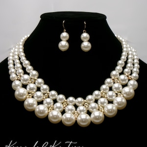May include: A three-strand pearl necklace and matching earrings. The necklace is made of large, white pearls and gold beads. The earrings are simple and feature two pearls each.