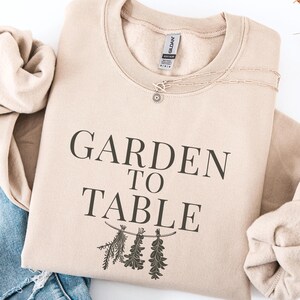 May include: A sand-coloured sweatshirt with the text "GARDEN TO TABLE" printed in black. Below the text is a botanical illustration. The sweatshirt is folded and placed next to a pair of blue jeans.