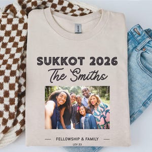 Może przedstawiać: Kremowa koszulka z napisem "SUKKOT 2026 The Smiths" nad zdjęciem rodzinnym. Poniżej zdjęcia widnieje napis "FELLOWSHIP & FAMILY LEV 23". Koszulka jest prezentowana z brązowo-białym swetrem w kratę i niebieskimi dżinsami.