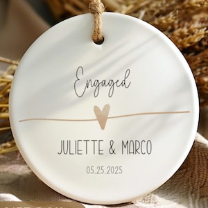 May include: A white ceramic engagement ornament with the word "Engaged" in script, a heart, and the names "Juliette & Marco" with the date "05.25.2025". The ornament hangs from a natural twine string.