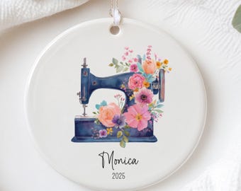 May include: A round, white ceramic ornament featuring a watercolor illustration of a blue sewing machine adorned with pink, peach, and purple flowers. The name "Monica" and the year "2025" are written below the sewing machine.