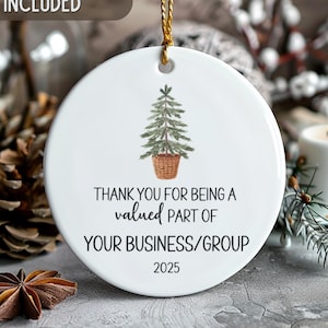May include: White ceramic Christmas ornament with a watercolor illustration of a Christmas tree in a basket. The ornament has the text "THANK YOU FOR BEING A valued PART OF YOUR BUSINESS/GROUP 2025". A gold string is attached for hanging.