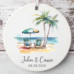May include: A round white ceramic ornament with a beach scene. The watercolor design features two palm trees, a beach umbrella, two beach chairs, and the text "John & Cassie" and the date "08.08.2025". A white ribbon is attached for hanging.