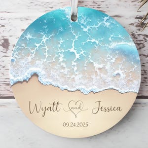 May include: Round ornament featuring a beach scene with turquoise water and tan sand. The ornament has the names "Wyatt and Jessica" with a heart symbol and the date "09.24.2025" printed on it. A white ribbon is attached for hanging.