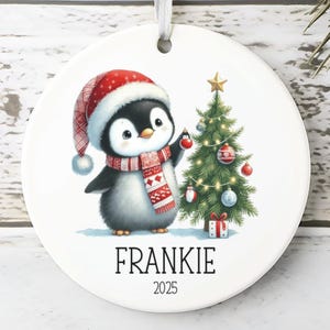 May include: A white ceramic Christmas ornament featuring a penguin wearing a Santa hat and scarf, decorating a Christmas tree. The ornament has the name "FRANKIE" and the year "2025" printed on it. A white ribbon is attached for hanging.