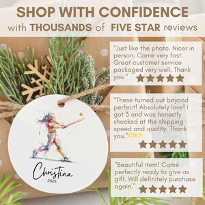 May include: A round, white ornament featuring a watercolor illustration of a softball player swinging a bat. The name "Christina" and the year "2025" are written below the image. The ornament is part of a gift set.