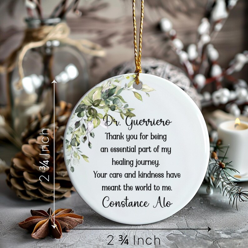 K&ouml;nnte beinhalten: Wei&szlig;er Keramikschmuck, 7 cm Durchmesser, mit floralem Design und goldener Aufh&auml;ngeschnur. Der Schmuck tr&auml;gt den Text "Dr. Guerriero, Danke, dass Sie ein wesentlicher Bestandteil meiner Heilungsreise sind."