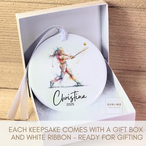 May include: White ceramic ornament with a watercolor illustration of a softball player swinging a bat. The ornament is personalized with the name "Christina" and the year "2025". It comes in a gift box with a white ribbon.