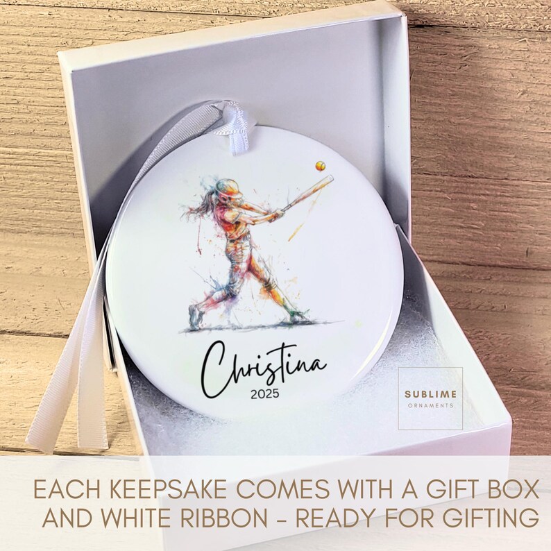 May include: White ceramic ornament with a watercolor illustration of a softball player swinging a bat. The ornament is personalized with the name "Christina" and the year "2025". It comes in a gift box with a white ribbon.
