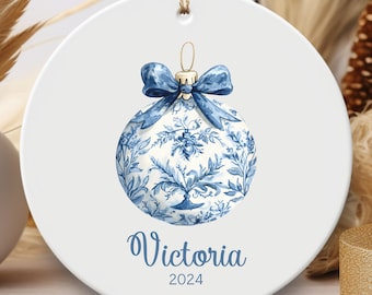 May include: A white ceramic ornament with a blue and white patterned Christmas ornament with a blue bow. The ornament has the name "Victoria" and the year "2024" written on it.
