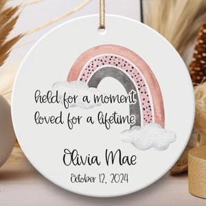 May include: A white ceramic ornament with a pink and gray watercolor rainbow, clouds, and the text "held for a moment loved for a lifetime". The ornament also includes the name "Olivia Mae" and the date "October 12, 2024".