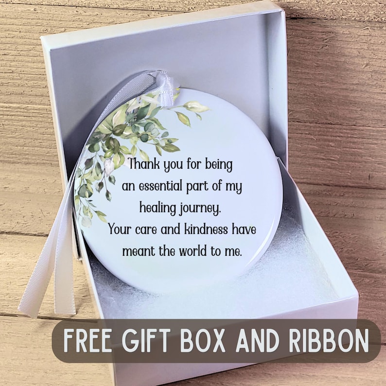 K&ouml;nnte beinhalten: Ein herzf&ouml;rmiger wei&szlig;er Ornament mit floralem Design und dem Text "Thank you for being an essential part of my healing journey." Es wird in einer wei&szlig;en Geschenkbox mit einem Band pr&auml;sentiert. Der Text "FREE GIFT BOX AND RIBBON" steht unten.