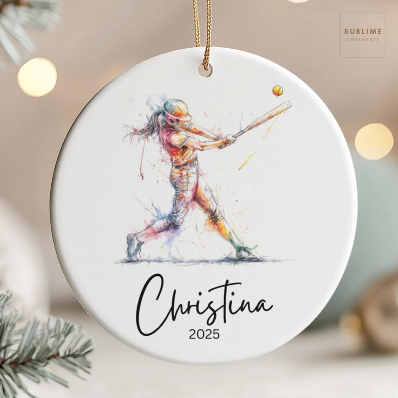 May include: White ceramic ornament featuring a watercolor illustration of a softball player swinging a bat. The design includes the name "Christina" and the year "2025" in black script. A gold string is attached for hanging. The ornament has the text "Sublime Ornaments" in the upper right corner.