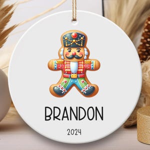 May include: White ceramic Christmas ornament with a gingerbread nutcracker design. The gingerbread man is wearing a red and blue uniform with a black hat. The ornament has the name "BRANDON" and the year "2024" printed on it.