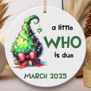 May include: A white ceramic ornament with a green Christmas tree with lights and a red bow. The text on the ornament reads "a little WHO is due MARCH 2025".
