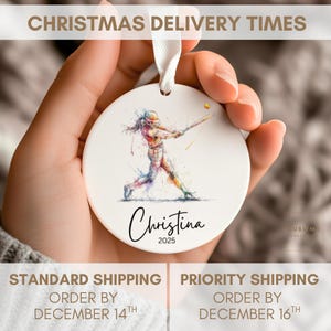 May include: White ceramic baseball ornament with a watercolor illustration of a batter swinging. The ornament has the name "Christina" and the year "2025" in a script font. A white ribbon is attached for hanging.