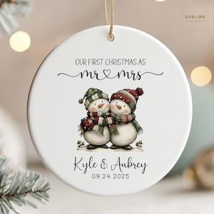 May include: A white ceramic Christmas ornament with a gold string. The ornament features two snowmen wearing hats and scarves. Text reads "OUR FIRST CHRISTMAS AS mr & mrs", with the names "Kyle & Aubrey" and the date "09.24.2025" below.