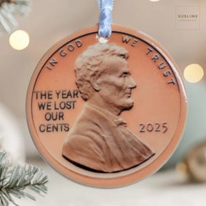 May include: A round, copper-colored Christmas ornament featuring a portrait of Abraham Lincoln. The ornament reads "IN GOD WE TRUST" and "THE YEAR WE LOST OUR CENTS 2025". It is suspended by a light blue ribbon.