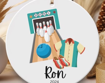 May include: A white round ceramic ornament with a bowling theme. It features a bowling alley with pins and a ball, a bowling shirt, and bowling shoes. The ornament is personalized with the name "Ron" and the year "2024".