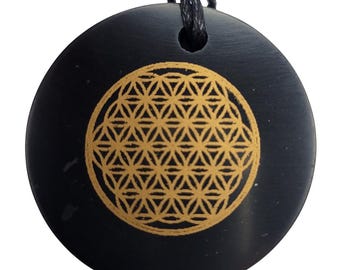 May include: A black, round pendant with a gold-colored Flower of Life design. The pendant is suspended from a black cord. The Flower of Life is a geometric pattern composed of overlapping circles.