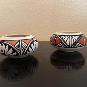 May include: Two small, round, ceramic bowls with intricate geometric designs. The bowls feature a white base, a band of orange, and black and white patterns. The bowls are displayed on a dark surface, reflecting their shapes.