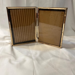 May include: A hinged, gold-toned metal picture frame, open to display two photos. The frame has a decorative edge and a brown backing. The glass is clear. The frame is resting on a white surface.