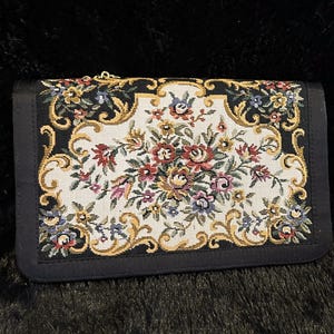 May include: A black clutch with a floral tapestry design. The tapestry features a cream background with a colorful arrangement of flowers in shades of red, pink, blue, and yellow, framed by gold and black borders.