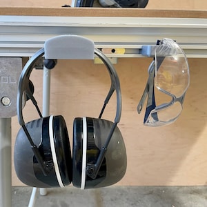 May include: A pair of black and gray earmuffs with white stripes and clear safety glasses hanging on a metal rail. The word "FESTOOL" is visible on a metal bracket.