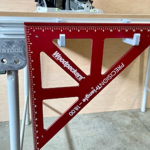May include: A red Woodpeckers 18.00 Precision Triangle with white markings and a white plastic hook on each side. The triangle is resting on a white table with a Festool label.