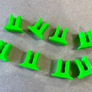 May include: Bright green plastic clips, arranged on a gray surface. Each clip has a T-shaped base with two prongs extending upwards, creating a U-shaped opening. The clips are evenly spaced and appear to be 3D-printed.