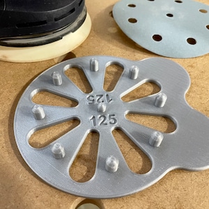 May include: A silver-colored sanding disc jig with 125 and 175 markings. The jig has a flower-like design with 10 holes around the center.