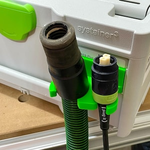 May include: A close-up of a green and black vacuum hose and electrical plug attached to a gray and white tool storage box. The plug is labeled "Plug it." The hose is held in place by a green clip.