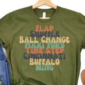 May include: Olive green t-shirt with a graphic design of the words "FLAP", "SHUFFLE", "BALL CHANGE", "MAKI FORD", "TIME STEP", "CINCINNATI", "BUFFALO", and "WING" in a retro font style.
