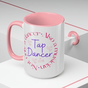 May include: White ceramic mug with a pink rim and handle. The mug has a pink circle with the text "Not only a dancer. Also a musician. Tap Dancer" in pink and purple text with pink stars.