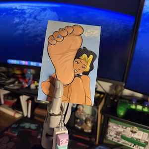 May include: A rectangular sticker featuring a cartoon illustration of a foot with blue nail polish and a woman with black and yellow hair. The sticker is held by a gray hand-shaped keychain with panda-faced cubes.
