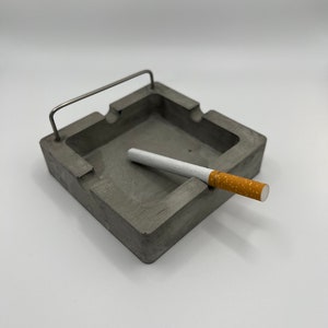 May include: A gray concrete ashtray with a metal handle and a cigarette resting on the edge.